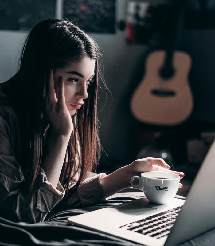 Woman lying on bed, focused on laptop with a coffee cup in hand, embracing remote work lifestyle.